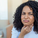 Žena s výraznou bolestí v krku si sahá na krk – angína, zánět krčních mandlí.